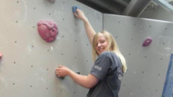A younger Abbi is wearing a grey T-shirt and rock climbing. She has paused to smile for the photo.