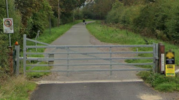 A large silver gate blocks the entrance to the bridleway, which is long and flanked by trees and bushes.