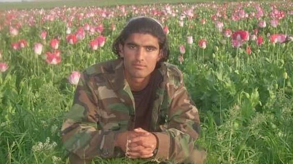 A young Wali Noori. He is crouching in a field of flowers with his fingers interlaced, wearing camouflage clothes.