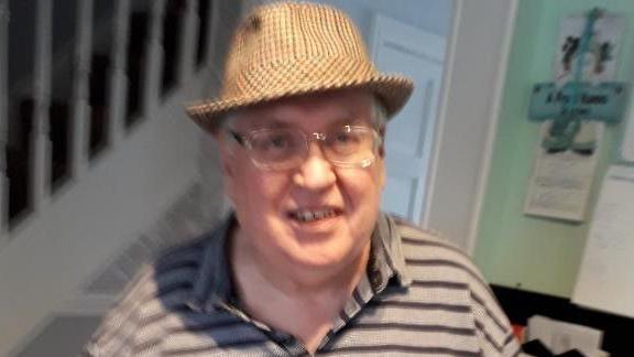 Mr Connolly is wearing a beige check hat on his head and transparent plastic spectacles on his face, a grey and black striped polo shirt with the top button undone. He is smiling into the camera.