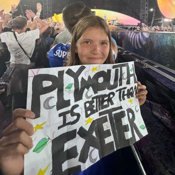 Lilly holding her sign which says 'Plymouth is better than Exeter'. She is smiling and is at the concert.