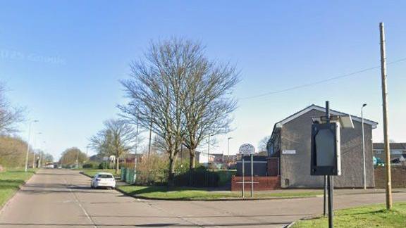 A suburban road junction with one white parked car ahead. Leafless trees, lampposts and a house are also visible.