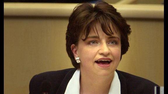 A young Wendy Alexander in 2000 in the debating chamber of the Scottish Parliament. She is sitting at a pale wood desk, and is wearing a white shirt and black jacket with a circular silver brooch.