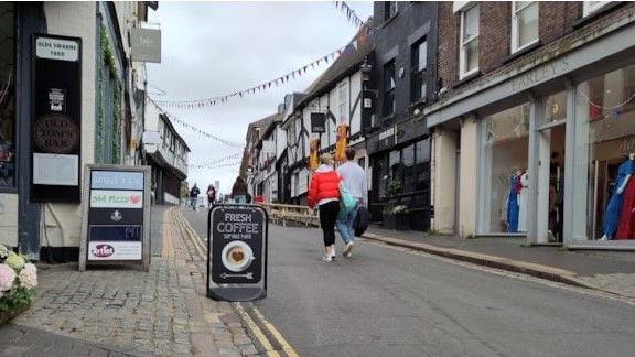 George Street, St Albans with shops on either side, some of which are Tudor styled white with black beams. Two people are walking in the road, one has a red jacket and black trousers and the other is wearing a grey jumper and blue jeans and is carrying a black bag.