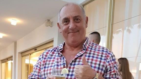 A man in a checked shirt smiling with his thumb up while holding a drink with a lemon slice