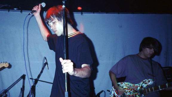 The Stone Roses during a live performance. Frontman Ian Brown, on the left, is wearing a navy t-shirt and is holding a mic up to the ceiling with his right hand and a mic stand with his left. On the right is bassist Mani. He is wearing a blue shirt and playing on a bass guitar.