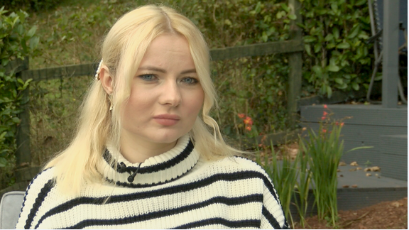 A head-and-shoulders photograph of Katya Piavka, a refugee from Ukraine who is living in Paignton. Katya has long blonde hair and blue eyes. She is wearing a white jumper with broad black horizonal stripes. A radio microphone is pinned on her collar. She is sitting in a garden with some tall orange flowers and decking in the background.