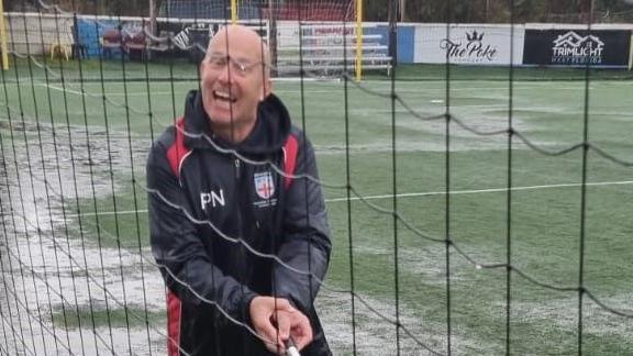 Paul Nicholls smiling as he sweeps water off an astro turf pitch. He is wearing a black jacket with his initials on.