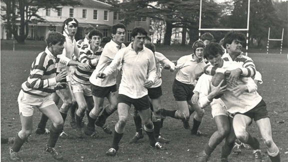 Black and white image of a group of men, some in white polo shirts and some in striped polo shirts, playing rugby union. One player has the rugby ball and the others are looking on as another player tackles him. 