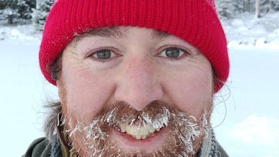 Oliver Eagle-Wilsher is smiling at the camera. He has a red beanie hat on. He is stood outside in the snow and has snowflakes on his ginger beard and hair. 