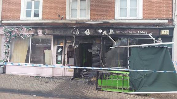 A burnt shop front, with broken windows, broken door, with police tape along the front. The signs to the shop are fire damaged, and items are outside on the pavement, as well as a green awning that has come down. A pink shop, next door, also has fire damage.