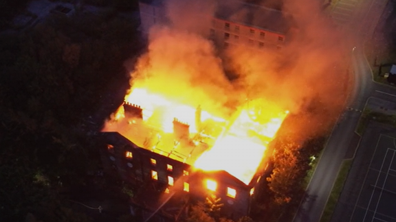 Image is an aerial shot of the fire raging at Woolton Hall. A ferocious orange glow emanates from within the building, which is otherwise shrouded in darkness.