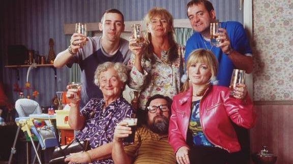 The Royle Family posed for a family photograph in their living room, holding drinks aloft. Standing backrow: Anthony, Barbara and Dave.  Front row seated: Norma, Jim and Denise.

A baby-chair is visible in the background