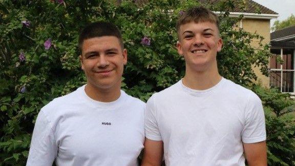 Two young men in white T-shirts smile at the camera as they stand side-by-side in front of a green hedge