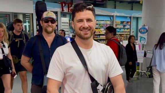 The image shows Ricky Eurys at an airport. He is smiling at the camera, and wears a white tshirt and a bag slung across his body.