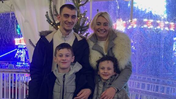 Jordan Williams, Natasha Goodall and their two children are at a christmas themed event. There is a Christmas tree in the background with neon lights coming from a fairground ride on the left.