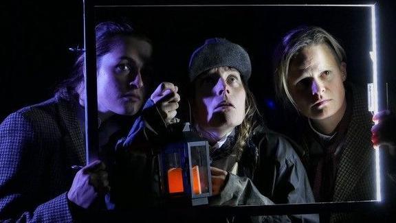 Three actors against a black background looking through a lit-up rectangular frame performing in a play about a Lowestoft-based 1970s paranormal magazine