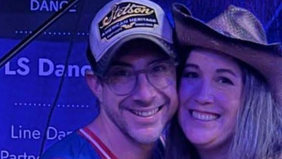 A man wearing a blue vest with a red trim, a black and white baseball cap and glasses. He has his arm around a woman in a black strappy top and jeans, who has long brown hair and is wearing a brown cowboy hat. They are both looking at the camera and smiling.
