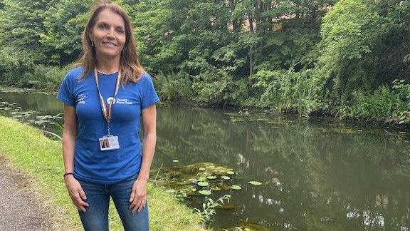 Henriette Breukelaar, who is wearing a blue Canal & River Trust T-shirt and jeans standing next to a canal, that has trees and shrubs on the other side