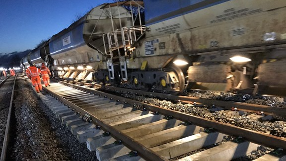 Nighttime on the railway line, can see a train track and a number of engineers wearing orange high vis boiler suits on the track