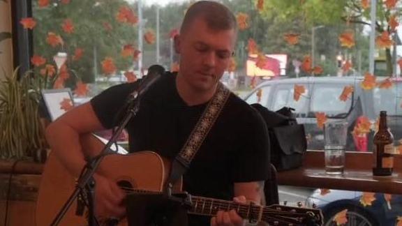 A young man with short hair and wearing a black t-shirt sits playing an acoustic guitar. A microphone on a stand is in front of him. He is inside a bar.