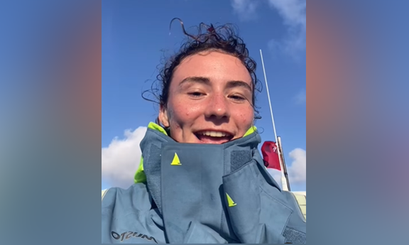 Zara Lachlan, whose dark hair is pulled back from her face. It is a close-up of her sitting in a boat in the Atlantic. She is wearing a water-proof coat and a purple fleece can be seen beneath. Behind her can just be glimpsed blue ocean on her right and above is blue sky