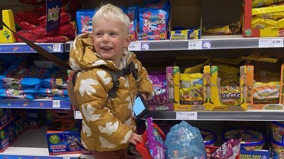 Oscar wearing a mustard coat pushing a child's shopping trolley with sweets in the basket in Asda.