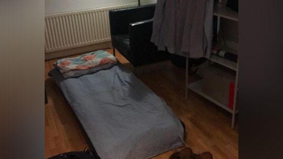 A blow-up mattress on the floor of a London flat. There are shoes and clothes hung up nearby.