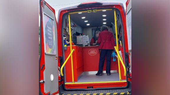 The back of the mobile post office van. The van's back doors are open, revealing a red, Post Office-branded counter. There is a member of staff serving a customer at the counter.