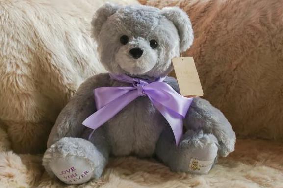 A grey teddy bear with a purple bow around its neck and "love you forever" embroidered on its foot.