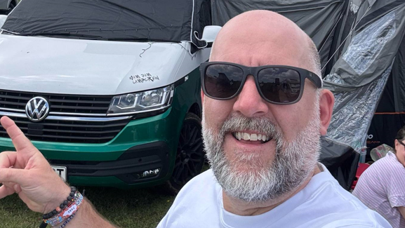 A selfie of Scott Siwicki. He is standing in front of a VW campervan and wearing dark sunglasses and a white T-shirt. He is smiling into the camera.