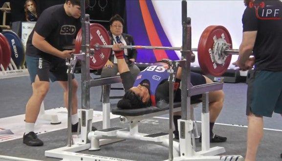 Siobhan Taylor bench pressing a bar, with red weights, accomponied by two spotters. 