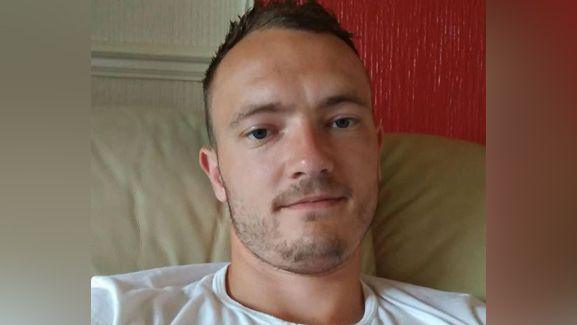 A man wearing a white T-shirt while sitting down. The photo is a selfie and he has gel in his hair and stubble 