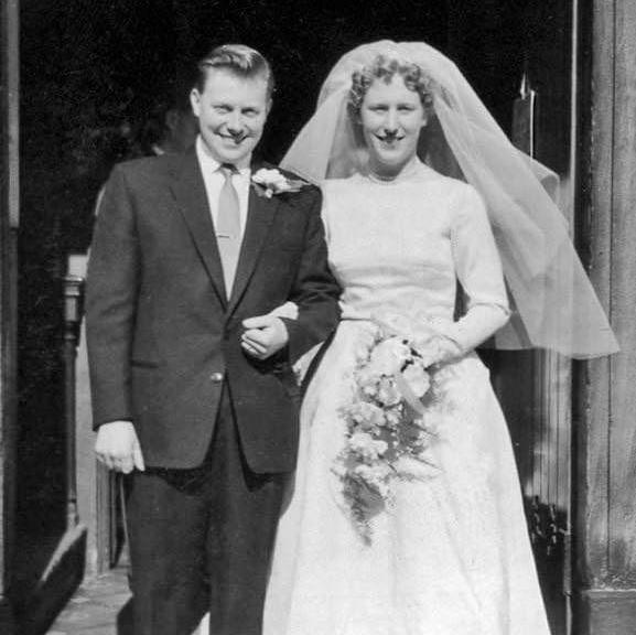 A black and white wedding photo from the 1960s showing a smiling man in a dark suit arm in arm with his bride in a white dress and veil carrying a bouquet