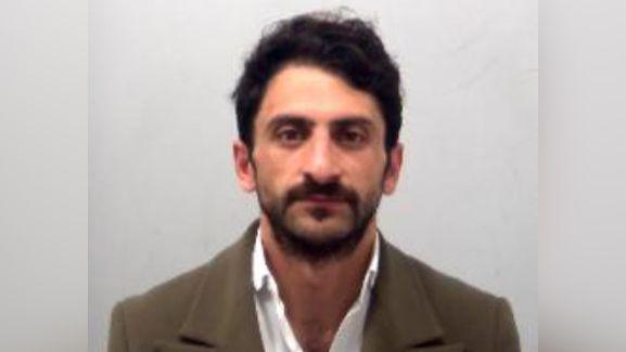 Ceyhan Dinler has a beard and black hair. He is wearing a white shirt and a brown blazer. He is not smiling and looking directly at the camera.