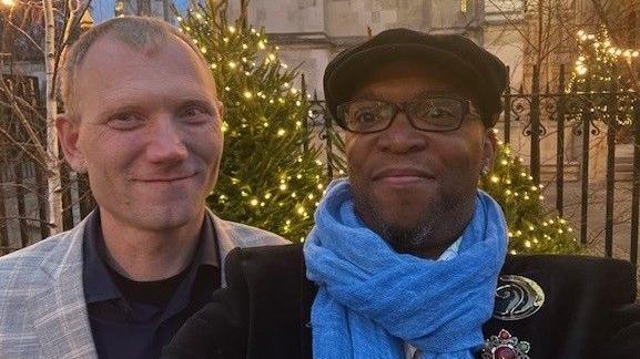 The image shows a couple smiling for a picture, the man on the left wearing a plaid blazer with a black shirt underneath, and his husband on the right is wearing a black coat with two golden broaches on his left collar, a bright blue scarf, glasses and hat. Behind them are trees with Christmas lights and a black fence in front of a beige building.