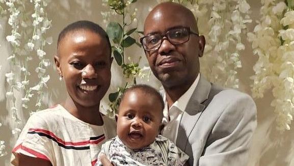 A smiling woman holds a baby next to a man in glasses. They all look at the camera