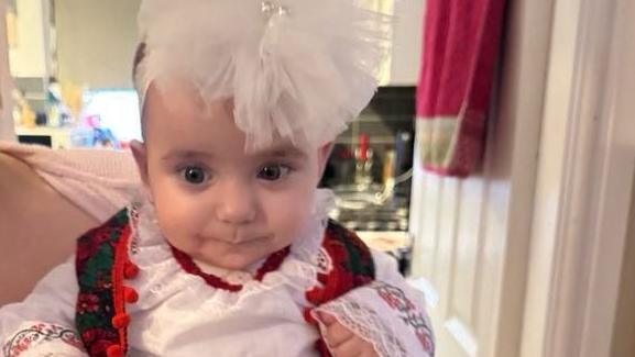 Baby Sophia Kelemen is wearing a white blouse/dress with embroidered detailing and a green waistcoat with red flowers and red embroidered edging. She is wearing a white ruffled hairband.