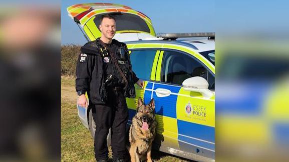A policeman standing next to a dog. The man is smiling and standing next to a police car.