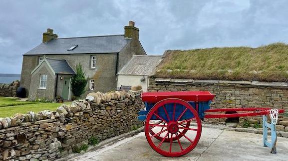 The farmhouse, seen from the outside, is a traditional built property with a small annex with a green front door. There is a turfed roof, stone-built farm building and drystone dyke that runs from the farm building to the foreground of the image. In a small enclosure is a red and blue painted horse-drawn cart.