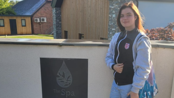 Catherine Blackhurst standing in jeans and a black zipped top under an open grey hoodie, with shoulder-length long dark hair and carrying a shoulder bag. She is standing in front of a sign that says 'The Spa'