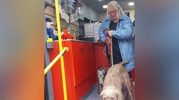 A woman and with two dogs - one a greyhound, the other possibly a labrador - leaving the mobile Post Office. She has long, blonde hair and is wearing a blue padded coat and glasses. On the left of the picture is a red Post Office counter. A Postman Pat doll is sitting on the counter, which also has a display of cards pinned to a shelf.