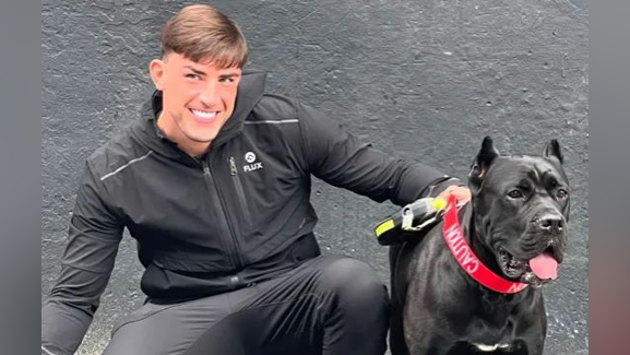 A man wearing a tracksuit smiling while holding on to the lead of his black cane corso