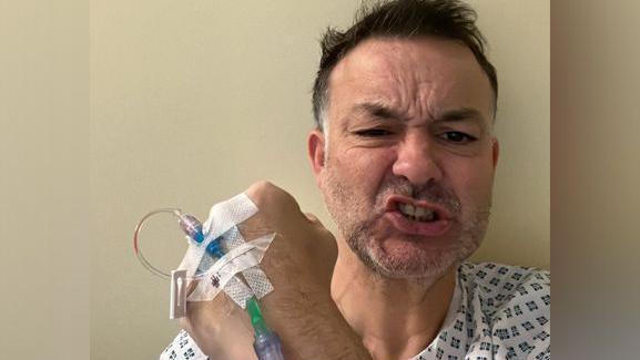 Lee Rawlinson is hospital holding his hand to the camera and pulling a face to show he is trying to be strong while undergoing treatment.