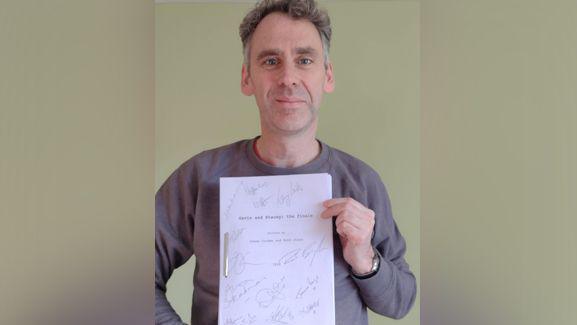 Robert Wilfort holding a signed copy of the script. He is smiling at the camera and wearing a grey sweatshirt.