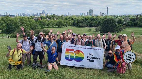 A group of people on a hill holding a banner that reads: "50 years of Gay Outdoor Club"