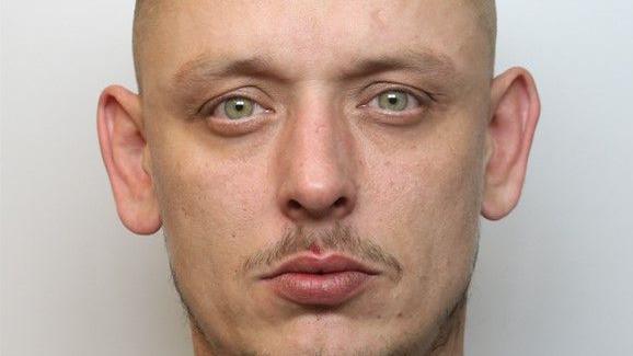 A police mugshot of a man with looking at the camera, with a moustache and hazel eyes.