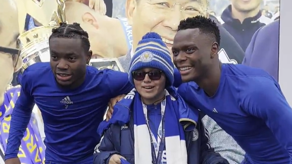 Two football players wearing blue undershirts with their arms around a boy, wearing a woolly hat and sunglasses