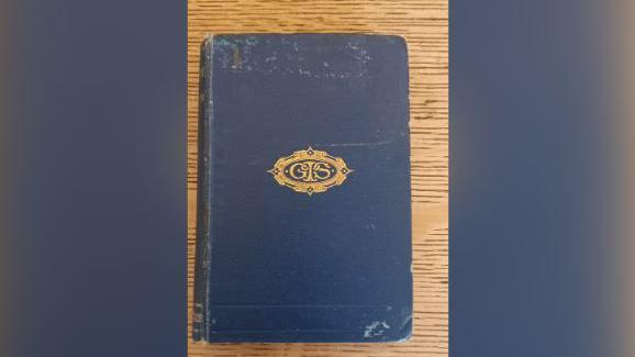 The book, which is closed and resting on a wooden table. It has a blue cover with a golden emblem printed in the middle with the initials GIS.