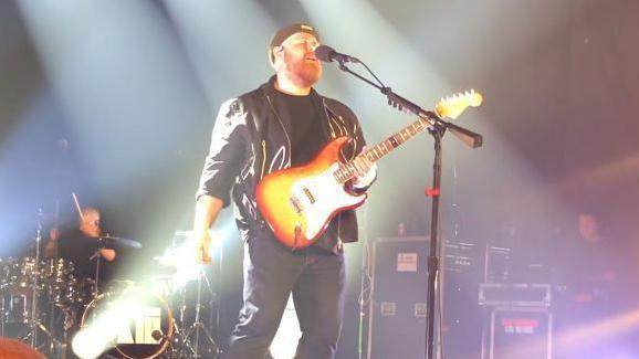 Tom Walker performing on stage, singing into a microphone on a stand as he holds a guitar